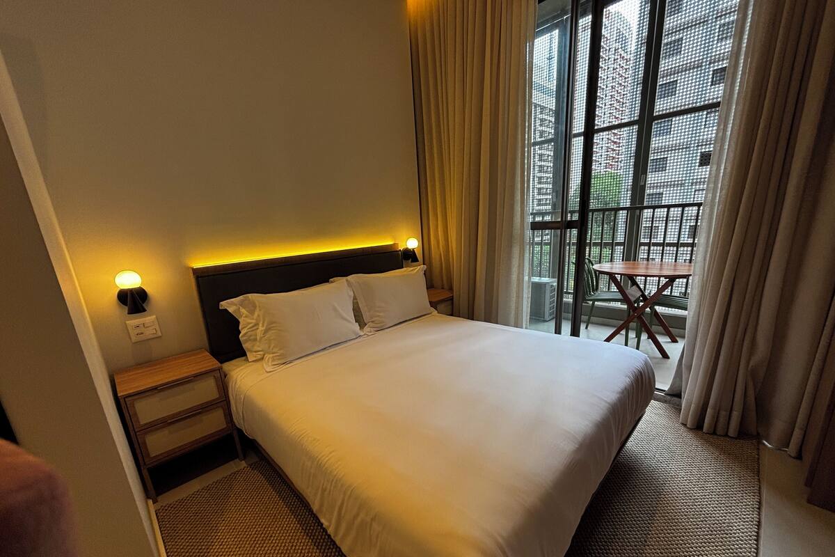 A hotel room with a bed, nightstands, a lamp, a window with curtains, and a view of the outside.