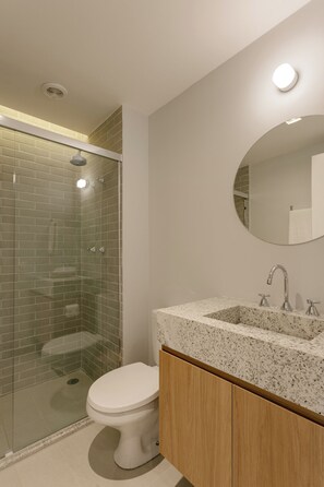 A bathroom with a glass-enclosed shower, a white toilet, a granite countertop sink, and a round mirror. - Aloe Casa Jardins (São Paulo)