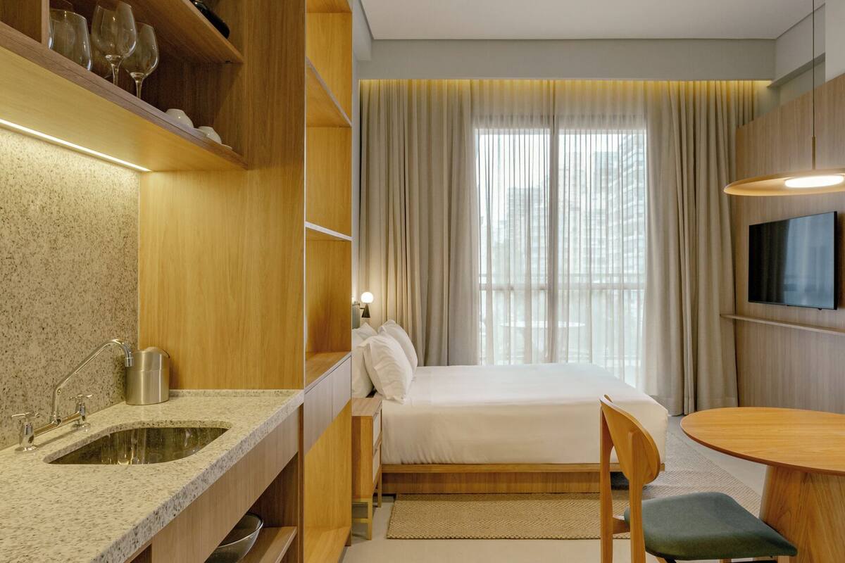 A modern hotel room with a kitchen area, a bed, a dining table, and a TV.
