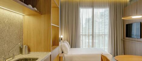 A modern hotel room with a kitchen area, a bed, a dining table, and a TV.