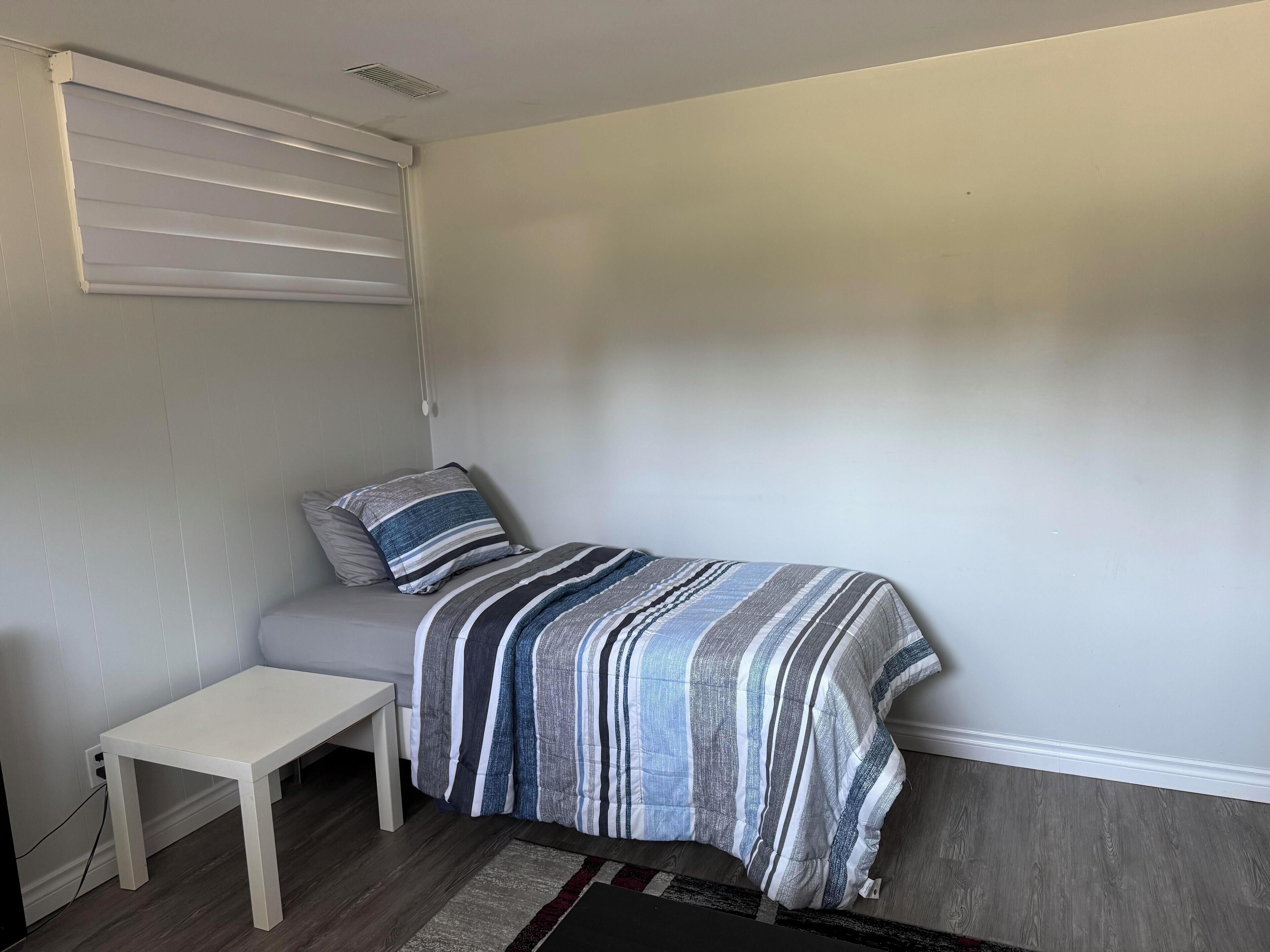 A small, neatly arranged bedroom with a single bed, a bedside table, and a window with blinds.