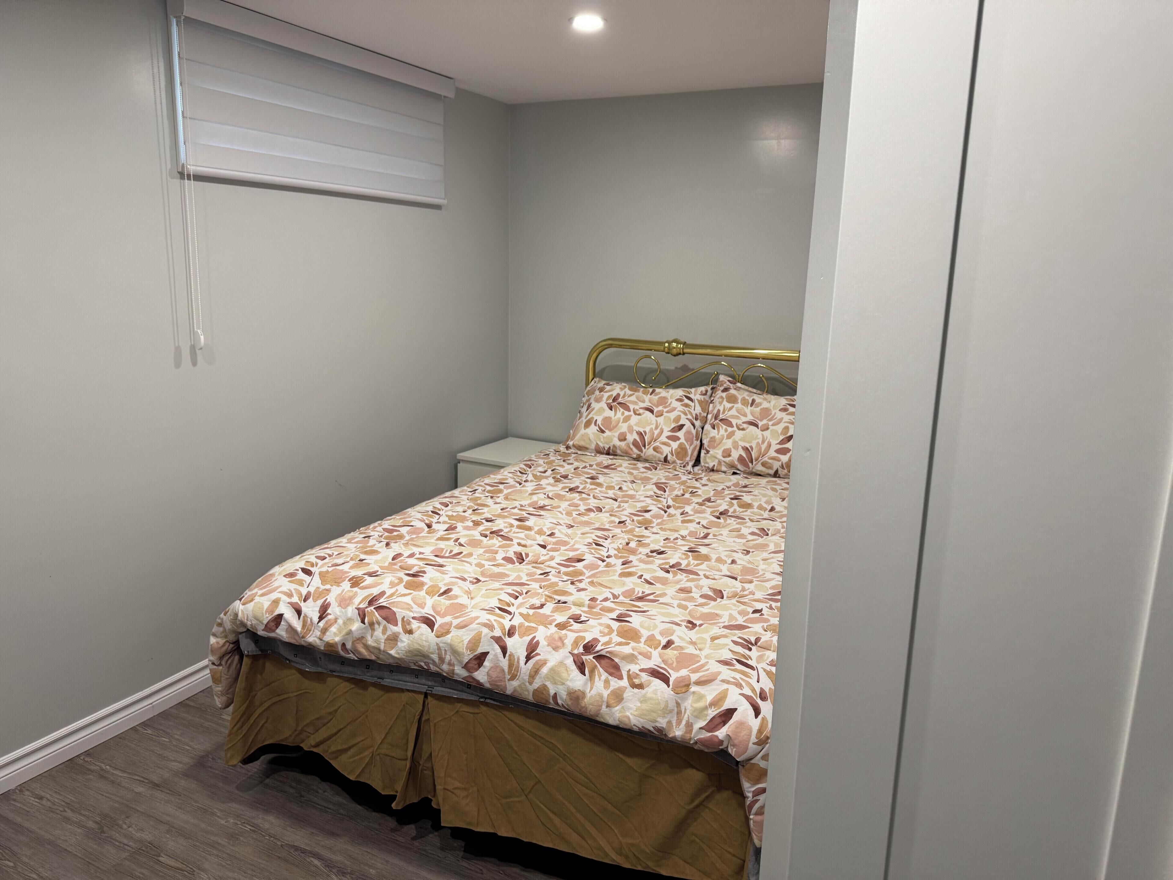 A bedroom with a bed, a nightstand, a window with blinds, and a wooden floor.