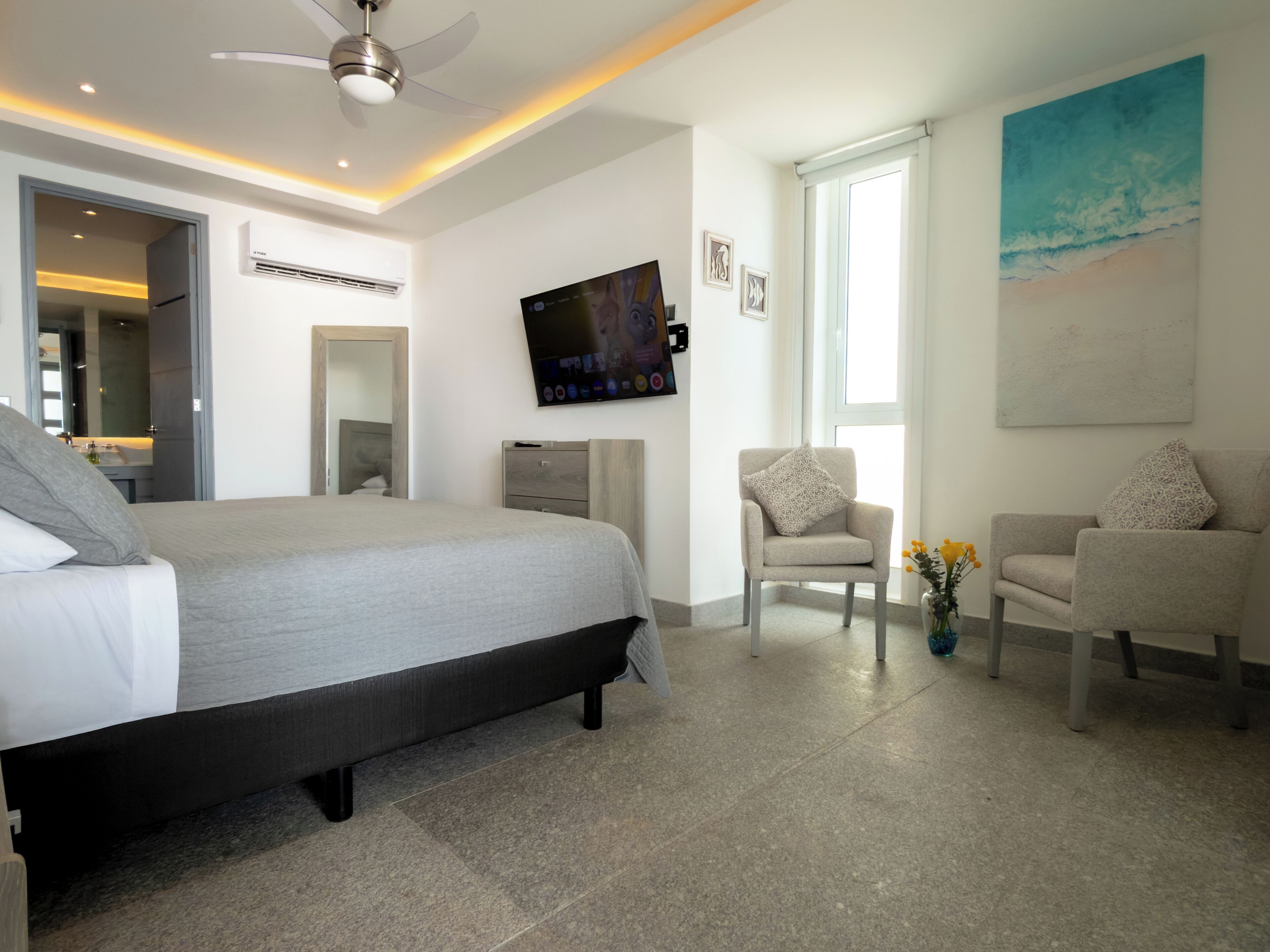 A modern bedroom with a large bed, a TV, two armchairs, and a painting of a beach scene.