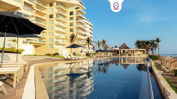 A resort pool area with a clear view of a multi-story building, lounge chairs, umbrellas, and a beach in the background. - Depto. Frente al Mar. Acapulco 4 Rec (Acapulco)