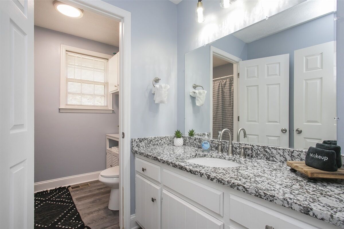 Une salle de bain avec un lavabo surmonté d’un plan de travail en granit, des meubles blancs, un grand miroir et un porte-serviettes.
