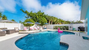 Una piscina con sillones de descanso y sombrillas, rodeada de palmeras y una valla blanca.