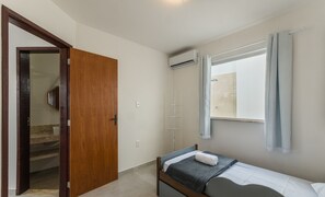A small, clean room with a single bed, a wooden door, a window with curtains, and an air conditioning unit. - Casa c\/ Piscina e 6 Quartos em Camaraçi PJE0001 (Camaçari)