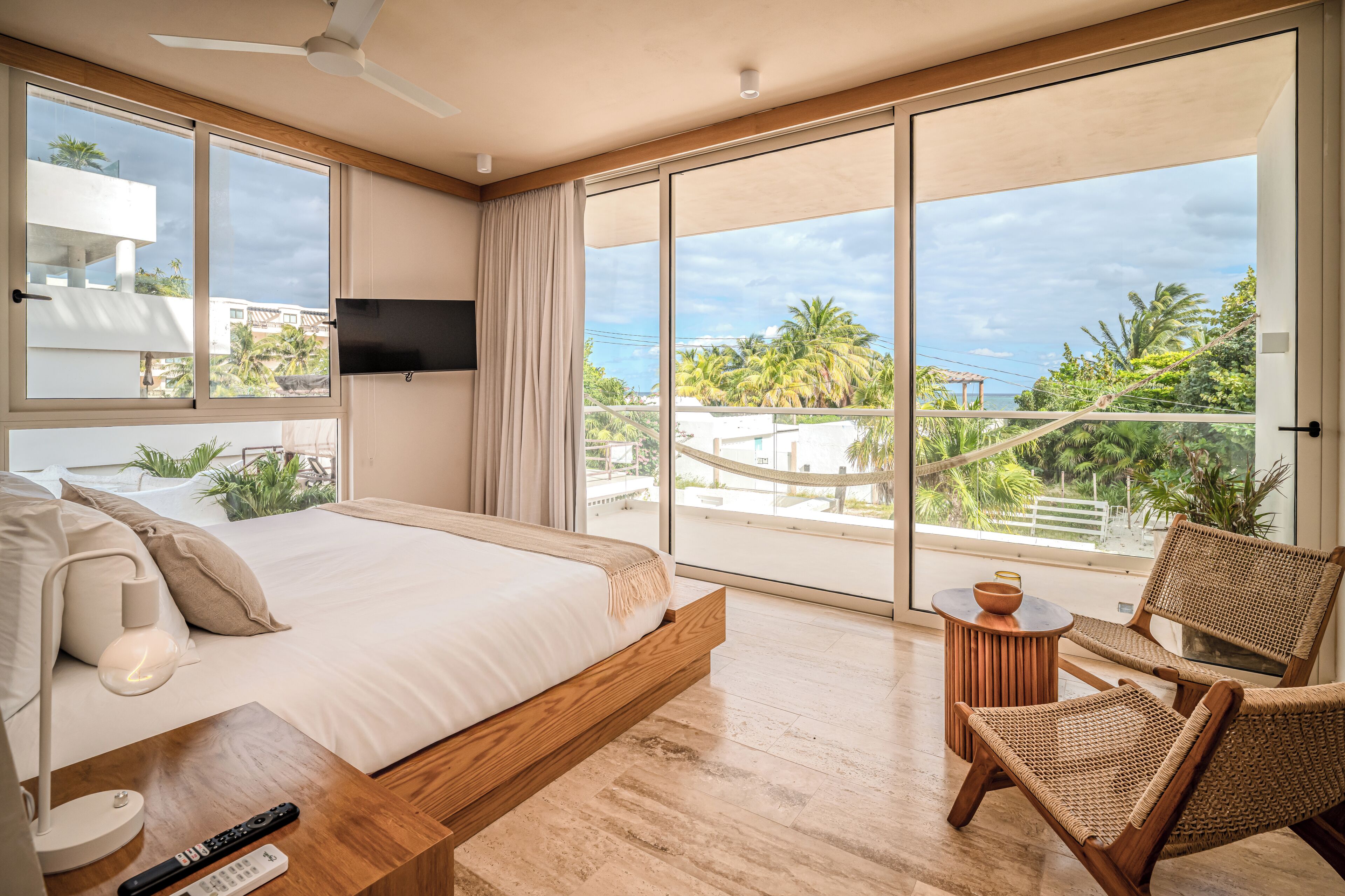 Quarto moderno com uma cama grande, uma televisão fixada na parede, uma varanda com vista para palmeiras e edifícios.