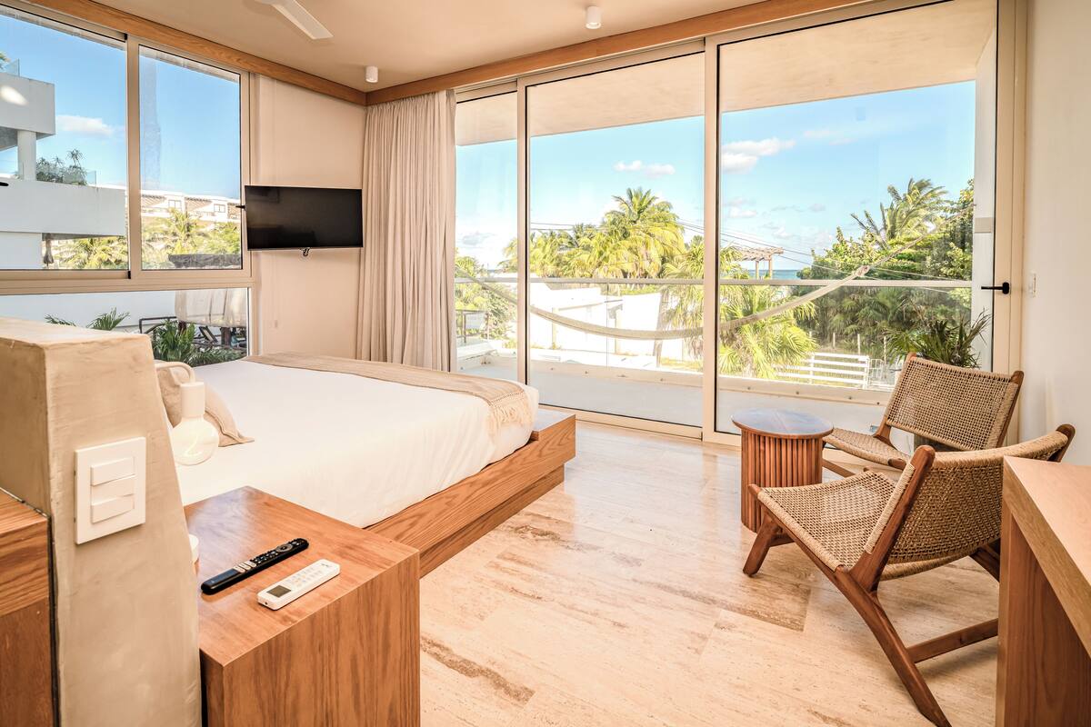 A modern hotel room with a large bed, a TV mounted on the wall, a balcony with a view of palm trees and buildings, and a wooden desk with a remote control.