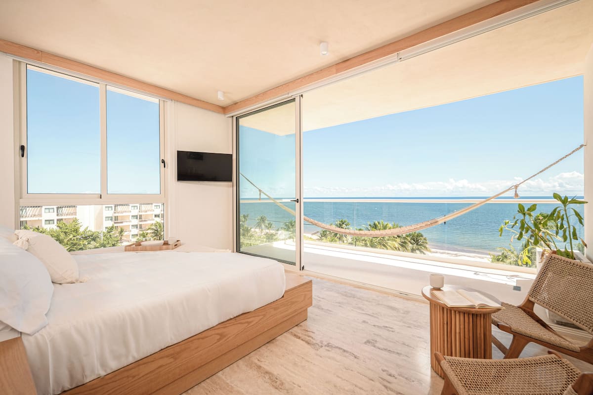 A bedroom with a large bed, a TV, a balcony with a hammock, and a view of the ocean.