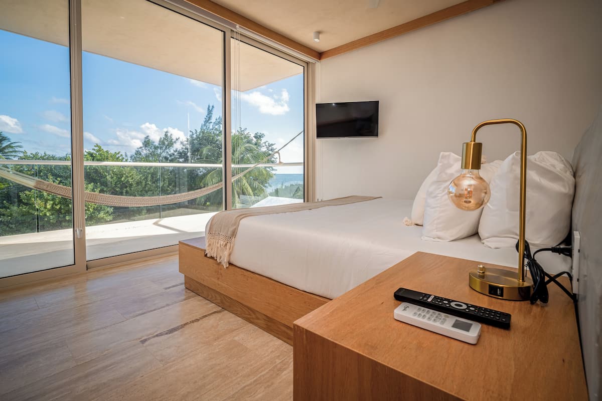 A modern bedroom with a large bed, a TV mounted on the wall, and a view of the outdoors through large windows.