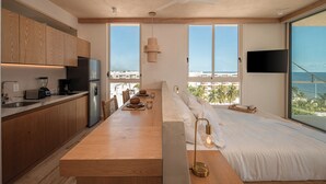 Una cocina moderna con armarios de madera, un refrigerador integrado y un comedor con mesa y sillas de madera. Se aprecia un amplio ventanal con vista al océano y un ventilador de techo.