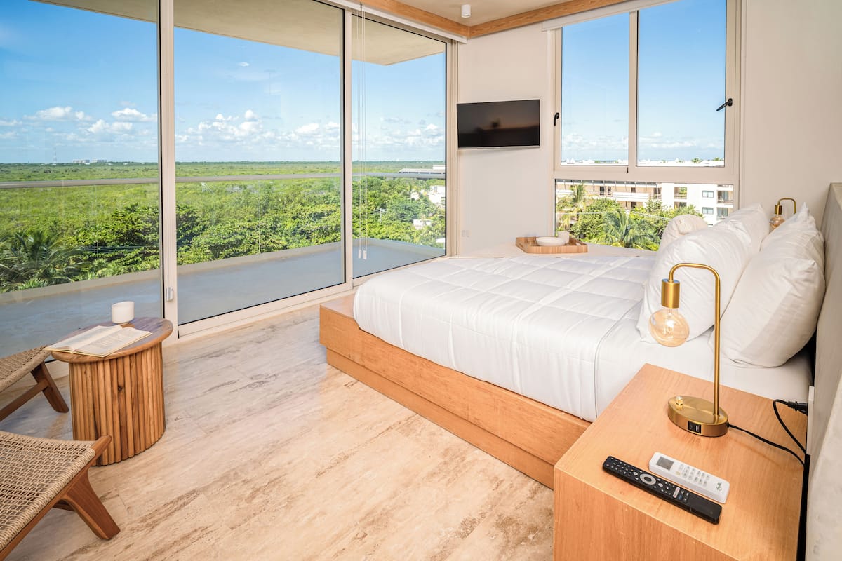 A modern bedroom with a large bed, a TV mounted on the wall, and a view of greenery outside.