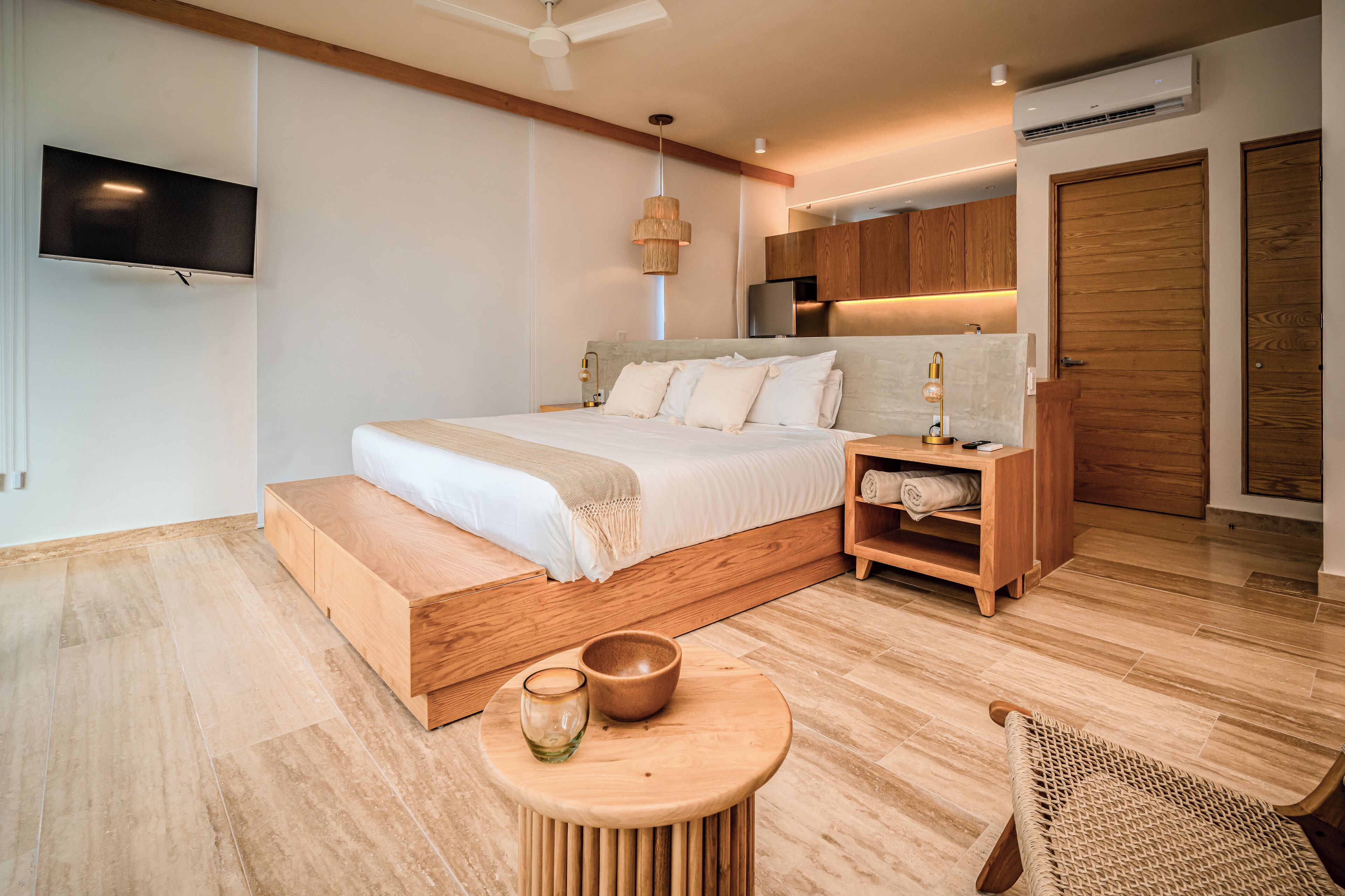 A modern bedroom with a wooden bed, a TV mounted on the wall, a bedside table with a lamp, and a small wooden cabinet.