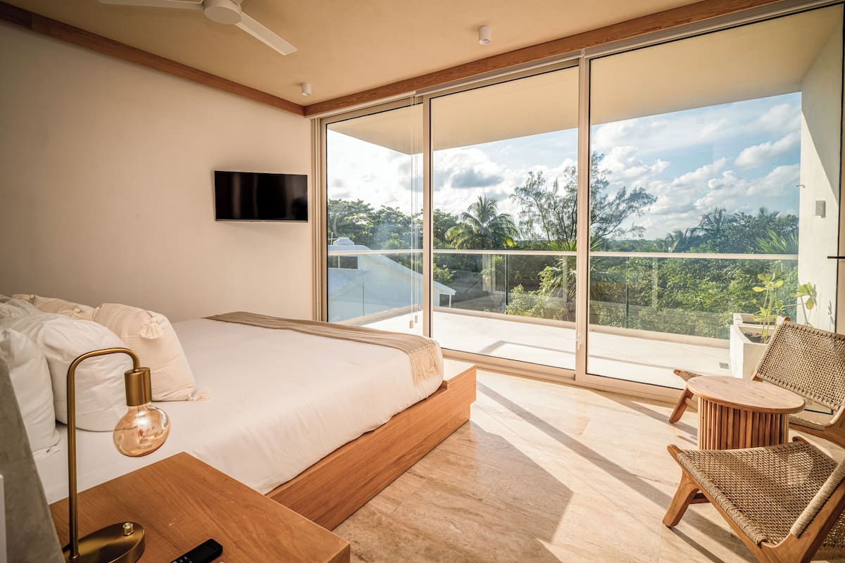 A modern bedroom with a large bed, a TV mounted on the wall, and a balcony with a view of the outdoors.