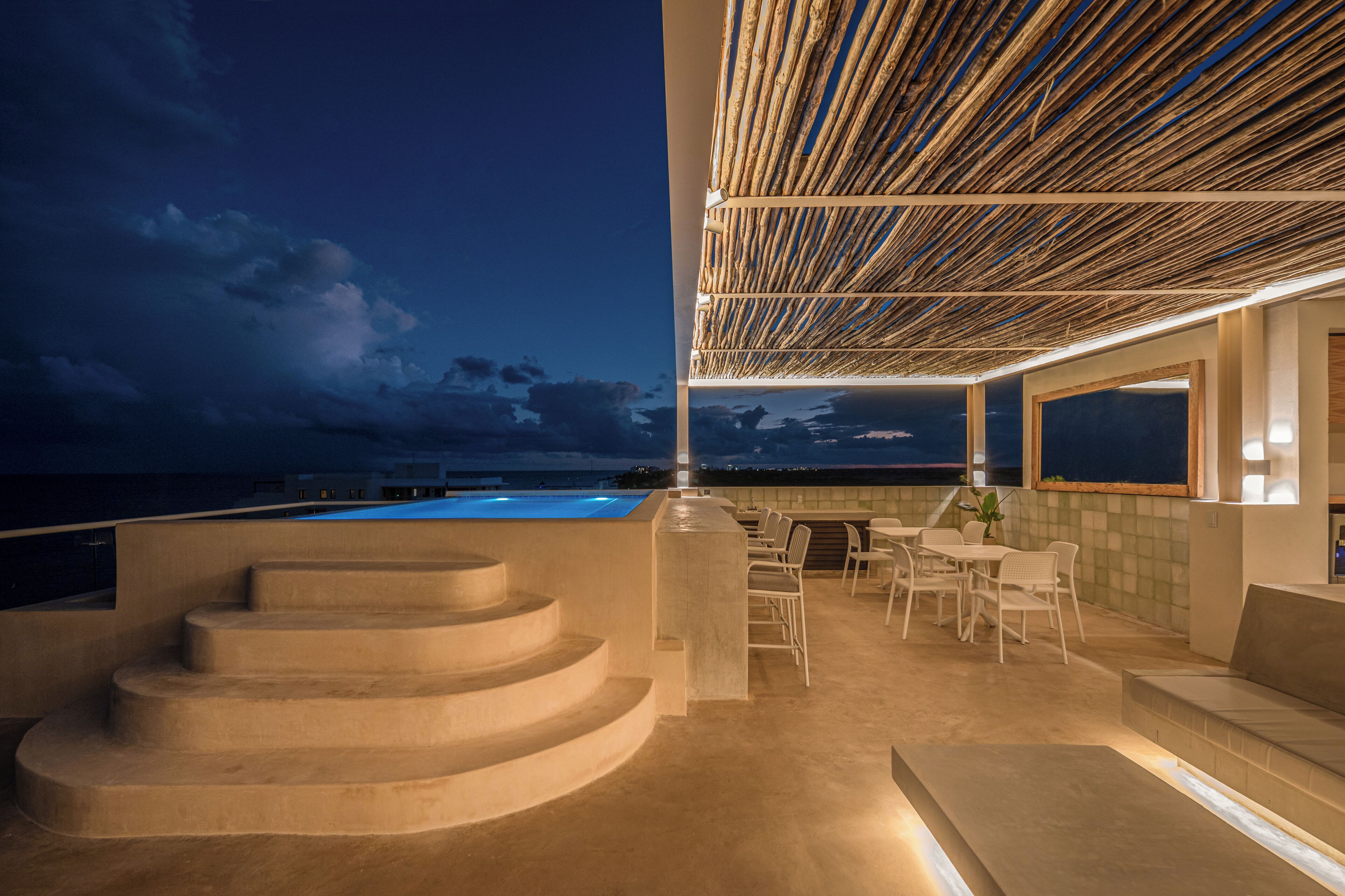 Terraza en la azotea con piscina, mobiliario exterior y vista al océano al anochecer.