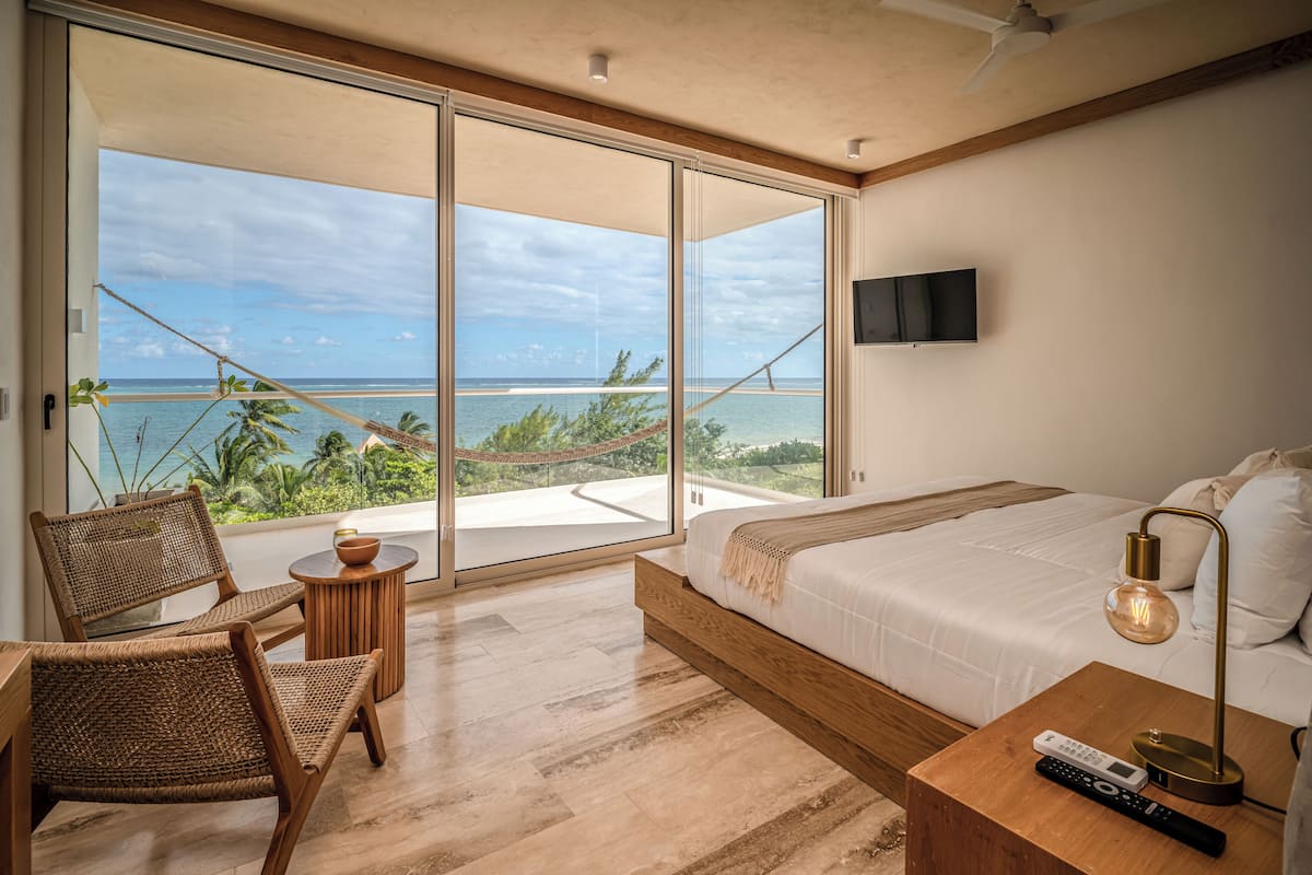 A modern hotel room with a large sliding glass door leading to a balcony with a view of the ocean. There is a bed with a wooden headboard, a bedside table with a lamp, and a chair.