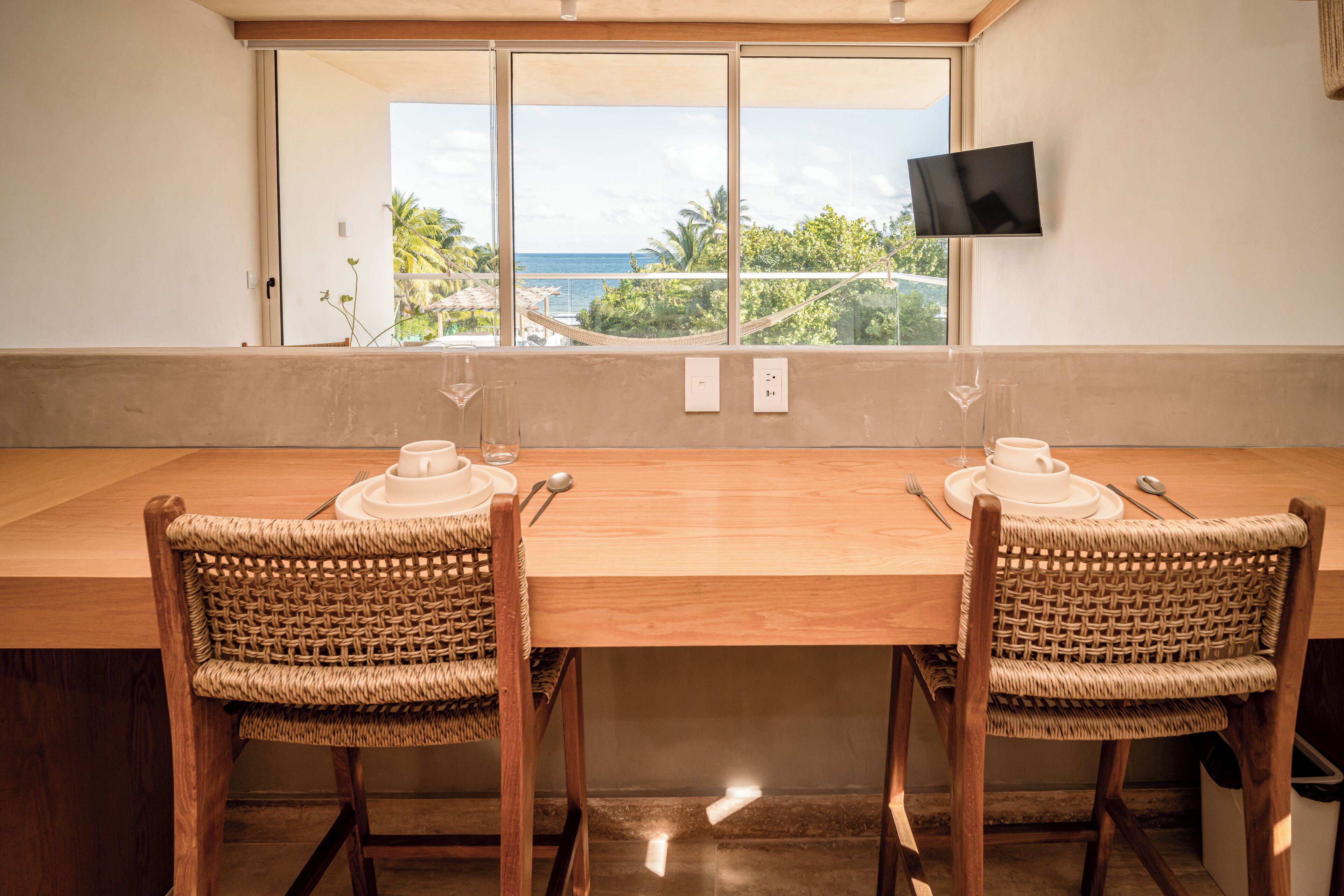 Un coin repas avec une table en bois et des chaises en osier, un téléviseur fixé au mur et une vue sur l’océan grâce à de grandes fenêtres.