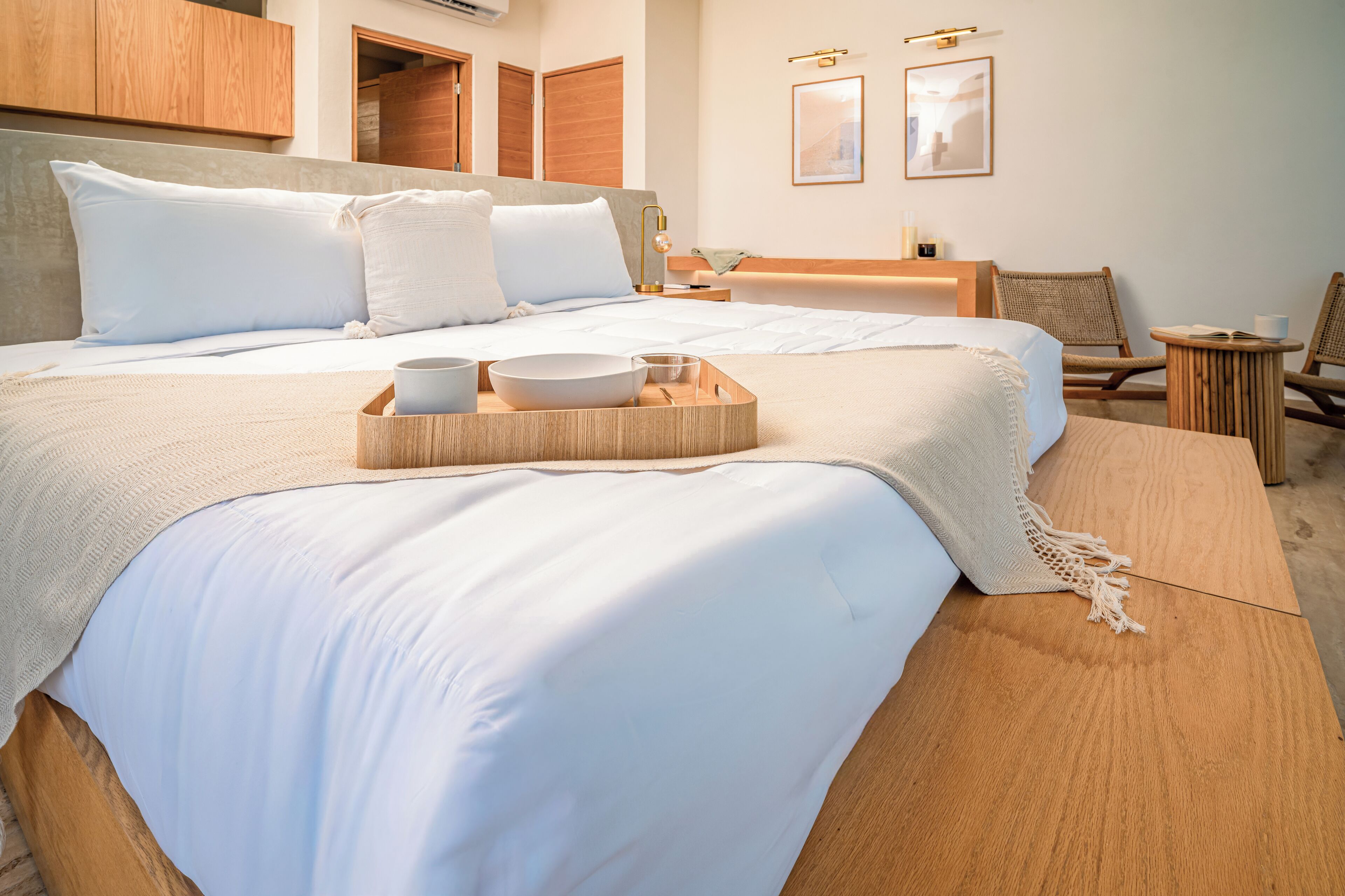 Una habitación de hotel con dos camas, cabecera de madera, mesita de noche con lámpara, una pequeña mesa redonda con bandeja y dos sillas de mimbre.