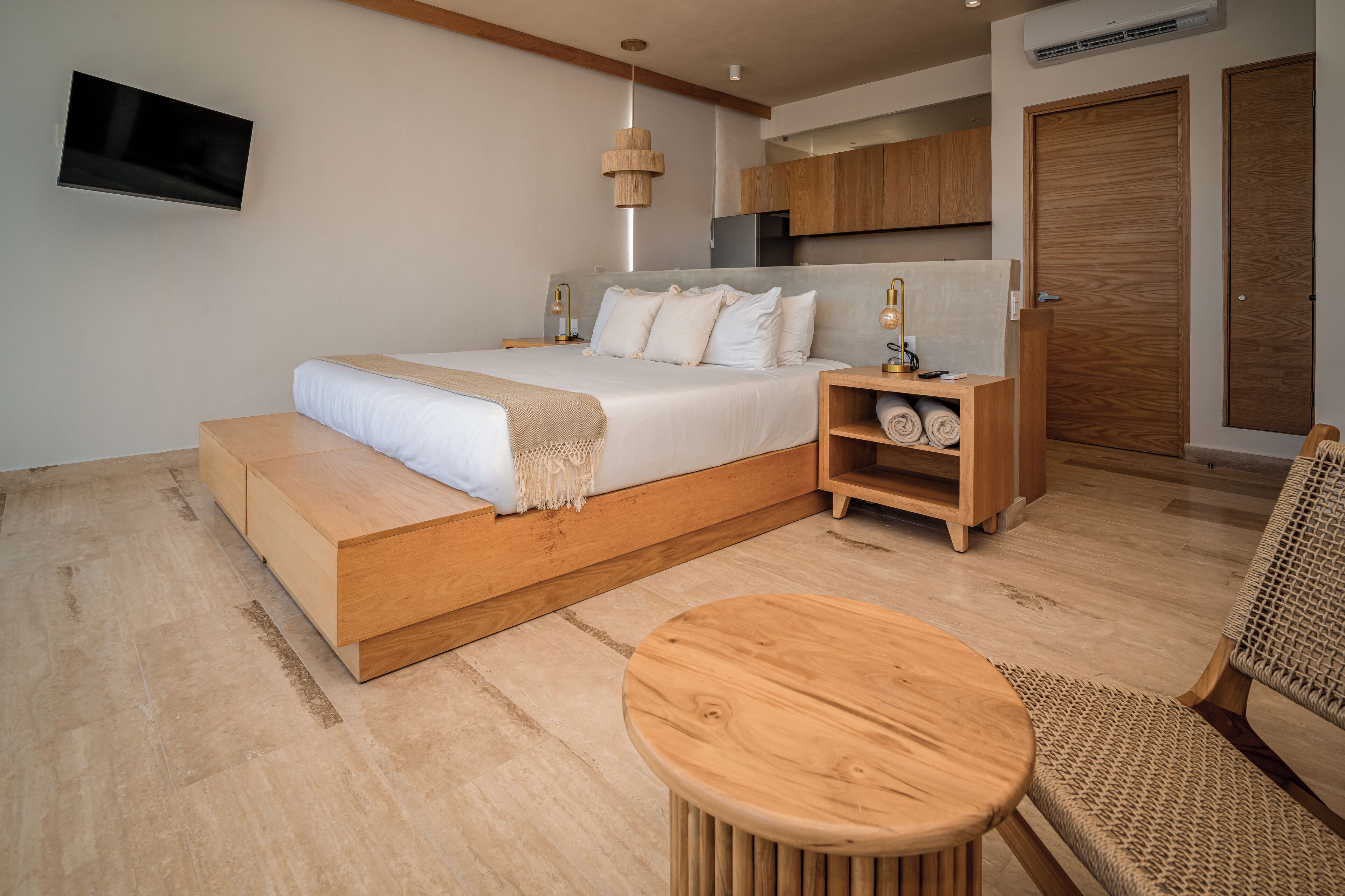 Un dormitorio moderno con una cama grande, cabecera de madera, mesitas de noche, una mesa redonda de madera y un televisor montado en la pared.