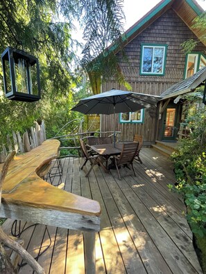 Um deck de madeira com mesa e cadeiras, cercado por vegetação e um edifício de madeira com janelas.