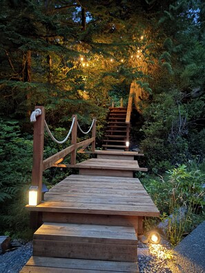 A wooden staircase leading up to a platform with a rope barrier, surrounded by dense greenery and string lights.