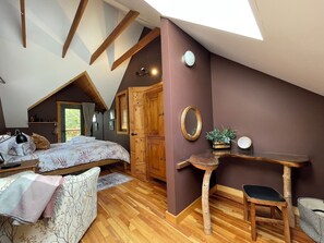 Um quarto de sótão aconchegante com teto inclinado, vigas de madeira, uma cama com colcha florida, uma escrivaninha com espelho e um pequeno banquinho.