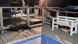 Une terrasse en bois avec un tapis bleu, des bancs en bois et un décor extérieur rustique agrémenté de guirlandes lumineuses.