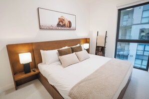 Ein Hotelzimmer mit einem großen Bett, einem Nachttisch mit Lampe, einem gerahmten Bild an der Wand und einem Fenster mit Blick auf Gebäude.