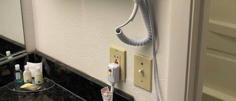 A bathroom with a hairdryer mounted on the wall, a sink with toiletries, and a towel rack with white towels.