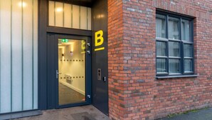 Une entrée d'immeuble avec une porte noire, un panneau jaune portant la lettre « B », et une fenêtre sur la droite.