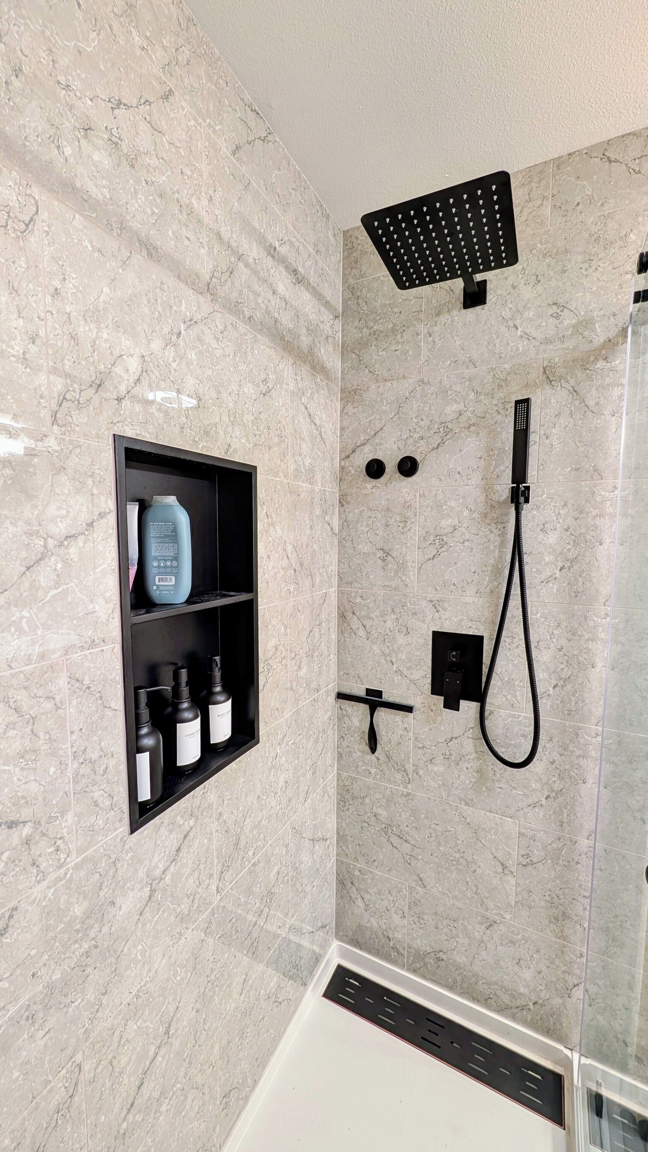 A modern bathroom with a marble wall, a built-in shelf with toiletries, a handheld showerhead, and a control panel.