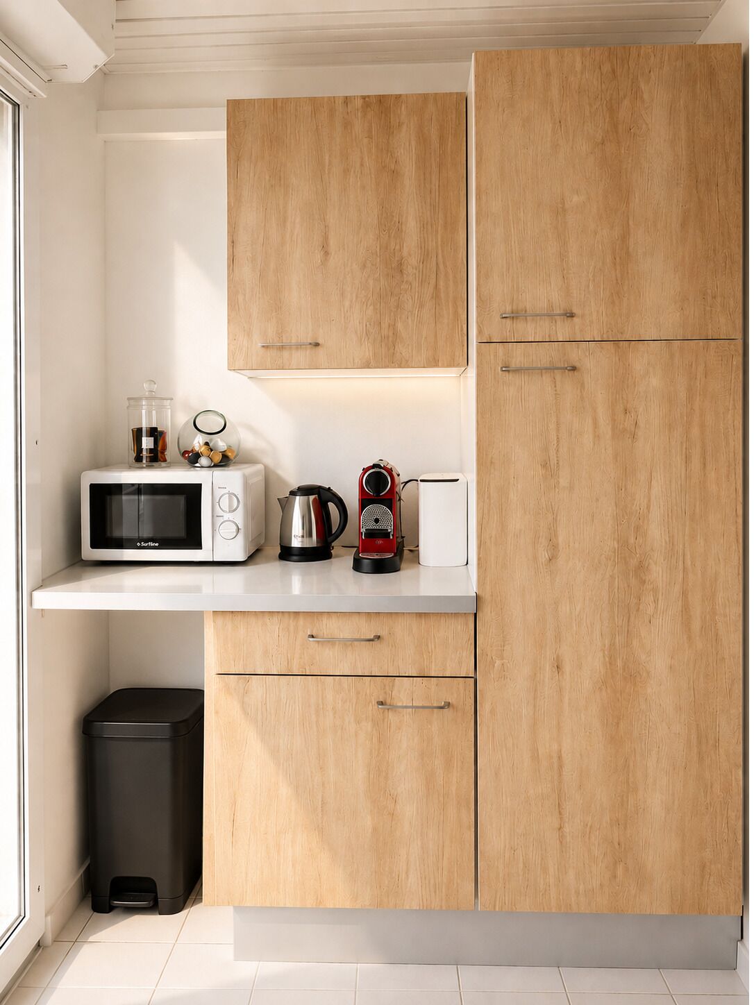 Una cucina compatta con forno a microonde, bollitore e macchina del caffè sul piano di lavoro, pensili in legno e cestino per i rifiuti.