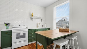 A kitchen with white tiled backsplash, green cabinets, a wooden table, and a window with blinds.