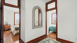 Un pasillo con suelo de madera, un espejo, una cama con colcha verde y una ventana con persianas.