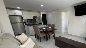 Een moderne woonkamer met een grijze bank, een eettafel met stoelen, een keuken met kasten en apparatuur, en een flatscreen televisie aan de muur.
