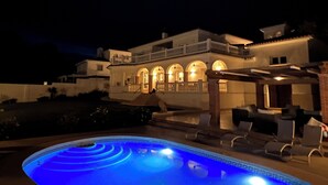 Una casa grande de dos pisos con piscina delantera, que cuenta con un porche cubierto con sillas de descanso.