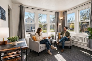 Una habitación con dos sofás enfrentados, una mesita con lámpara y ventanas grandes con cortinas.