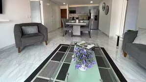 A modern living room with a grey sofa, a coffee table with a vase of flowers, and a dining area with a kitchen in the background. - Condominio de 2 dormitorios y 2 baños, piscina y parrilla en Cabo San Lucas (Cabo San Lucas)