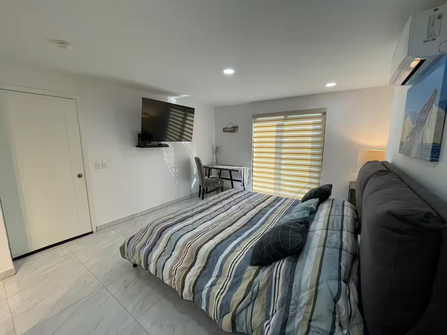 A bedroom with a bed, a desk, a chair, a TV, and a window with blinds.