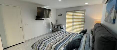 A bedroom with a bed, a desk, a chair, a TV, and a window with blinds.
