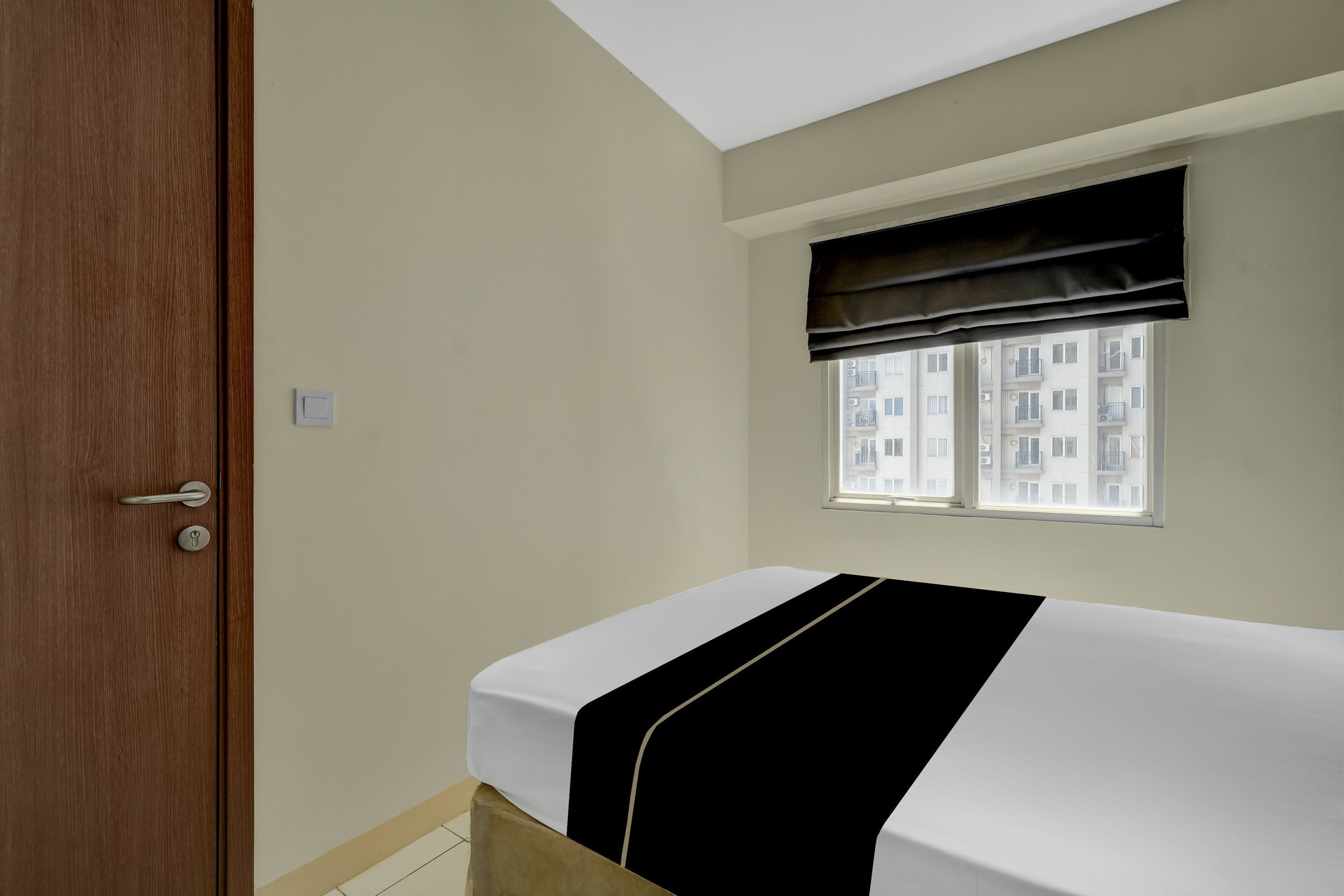 A room with a bed, a window with a black valance, a door, and a light switch on the wall.