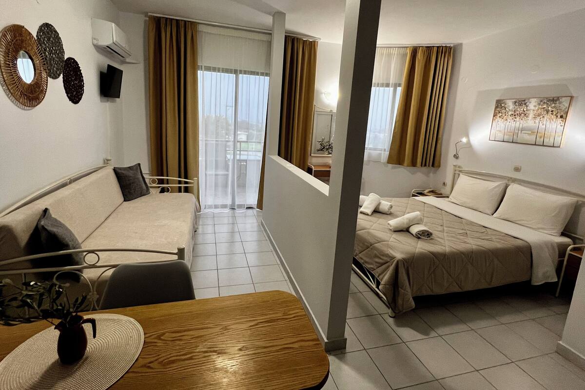 A hotel room with a bed, a sofa, a dining table with a vase, and a view of the outdoors.