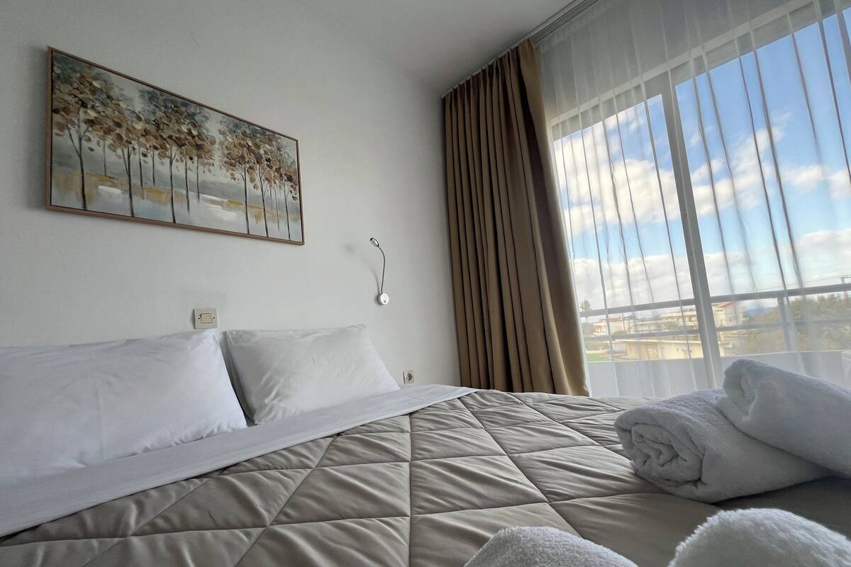 A hotel room with a bed, a painting, a window with curtains, and a view of the outside.