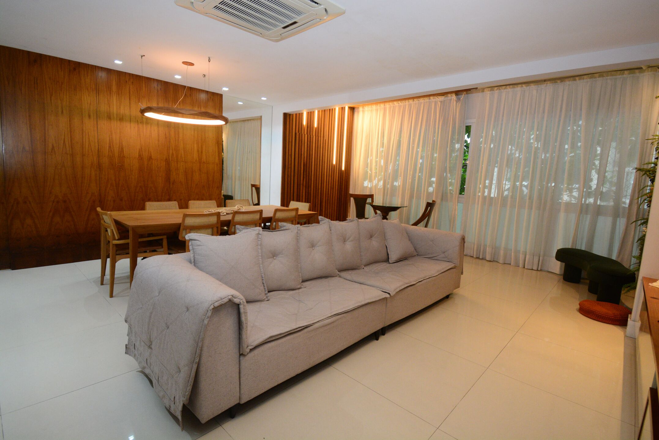 A modern living room with a beige sofa, wooden dining table, and chairs. There is a large window with sheer curtains and a plant on the right.