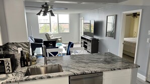 A modern kitchen with a granite countertop, stainless steel appliances, and a dishwasher. There is a dining area with a table and chairs, and a ceiling fan above. - Cozy 1-bedroom condo in peaceful Hallandale Beach with AC (Hallandale Beach)