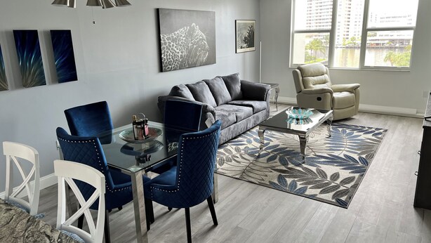 A modern living room with a gray sofa, blue chairs, a glass coffee table, and artwork on the walls. - Cozy 1-bedroom condo in peaceful Hallandale Beach with AC (Hallandale Beach)