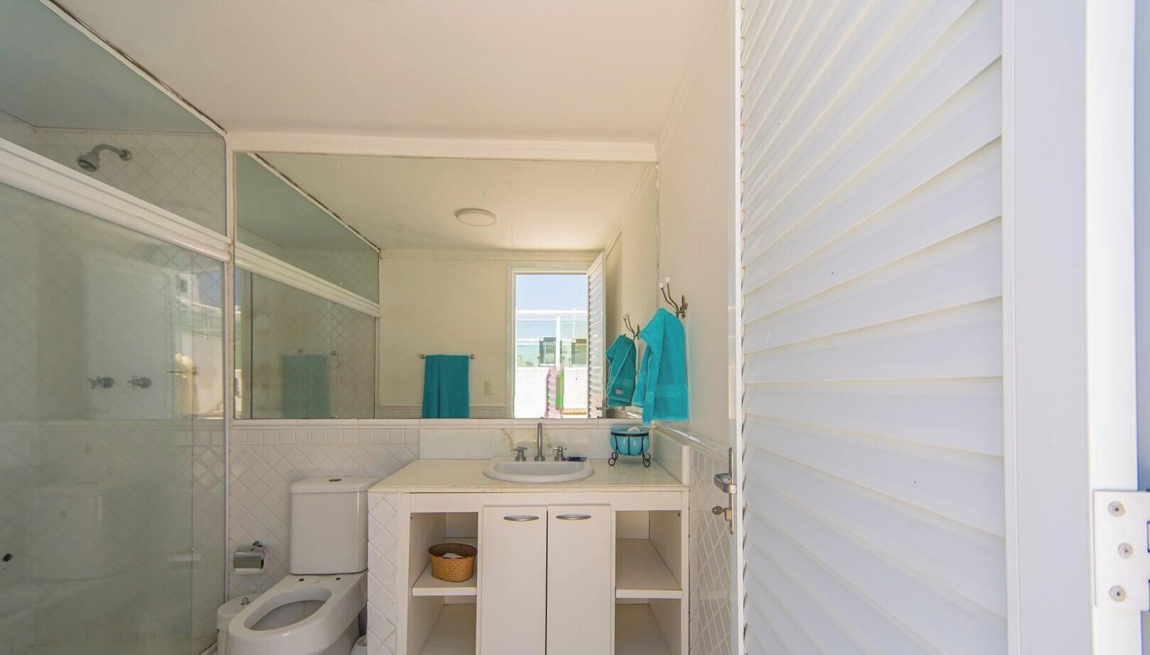 Une salle de bain équipée de toilettes, d’un lavabo et d’une douche. Des serviettes sont accrochées au mur et une fenêtre laisse entrer la lumière naturelle.