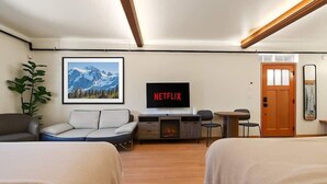 Ein Hotelzimmer mit zwei Betten, einem Schlafsofa, einem an der Wand montierten Fernseher, einem kleinen Tisch und einer Tür.