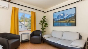 Ein Wohnzimmer mit einer Couch, zwei Sesseln, einem kleinen Tisch, einer Zimmerpflanze und einem großen Fenster mit gelben Vorhängen.